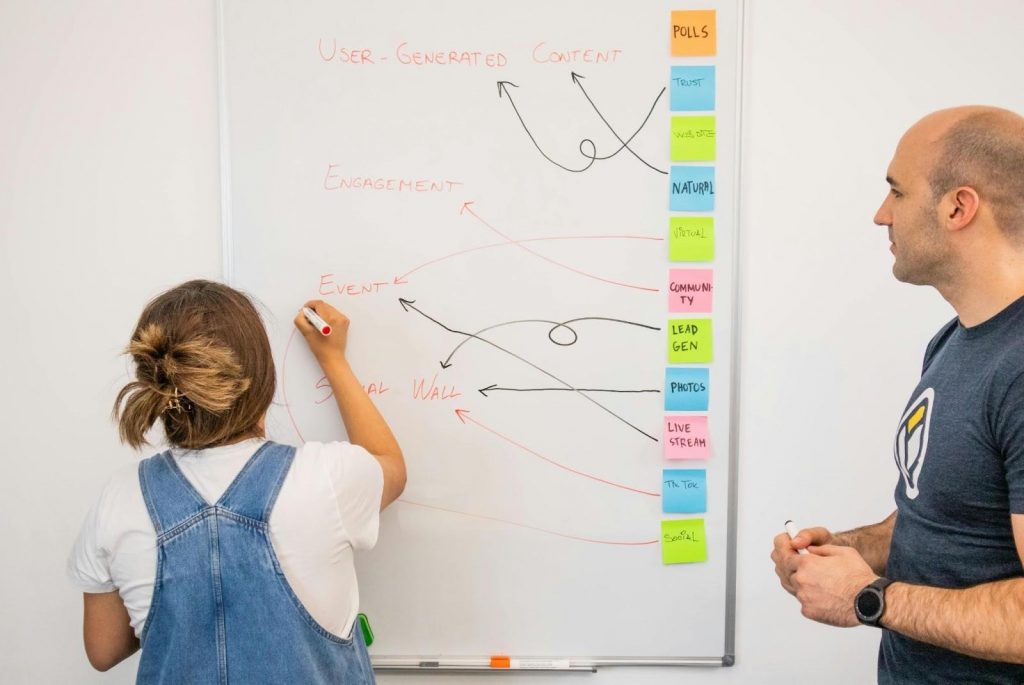 A woman writing events, user-generated content, and engagement on a whiteboard while a man observes, showing digital marketing for small businesses in action.
