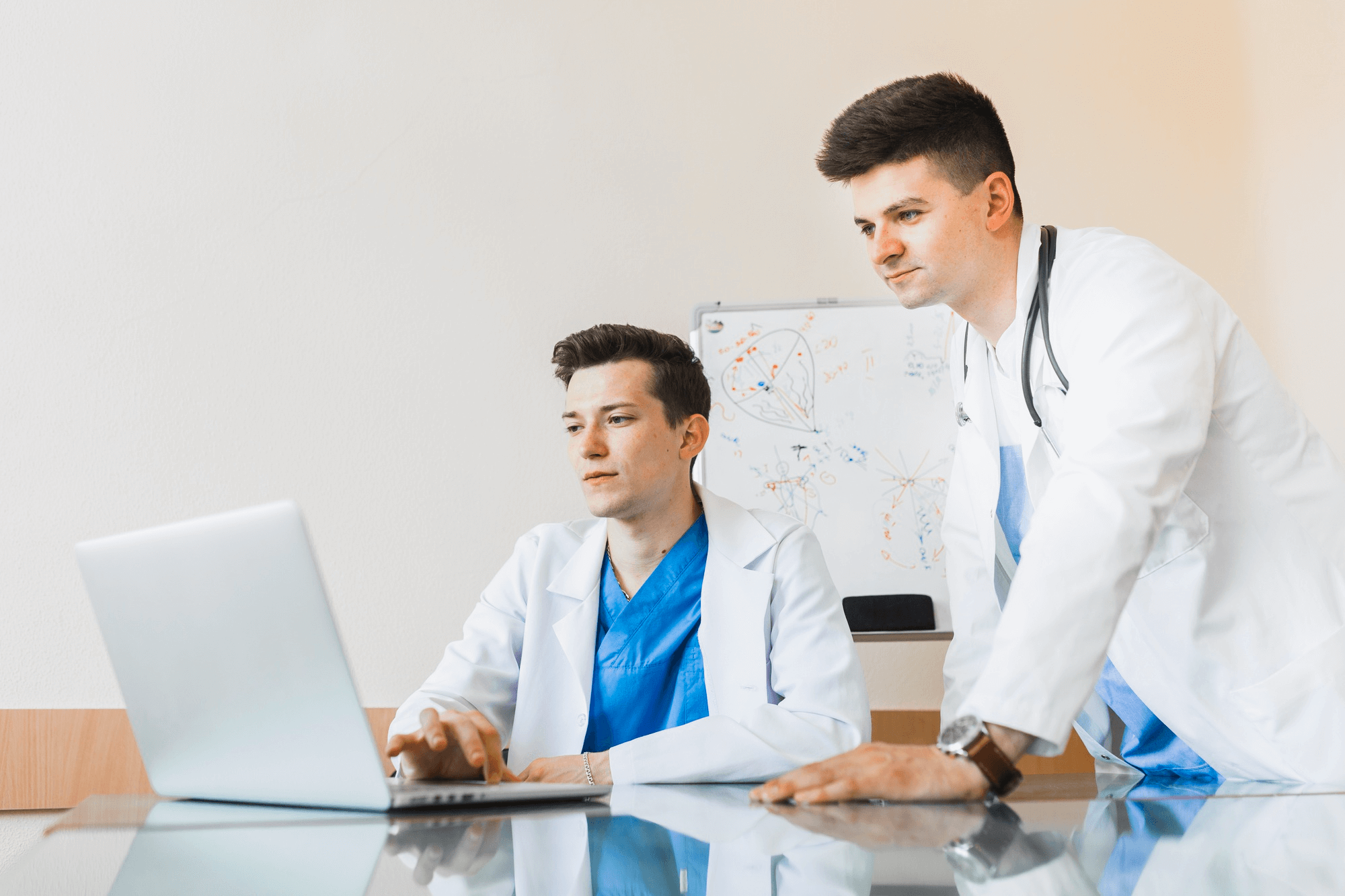 Two doctors in white coats focus on a laptop screen. One is seated typing, while the other stands beside him. A whiteboard with diagrams is behind them.