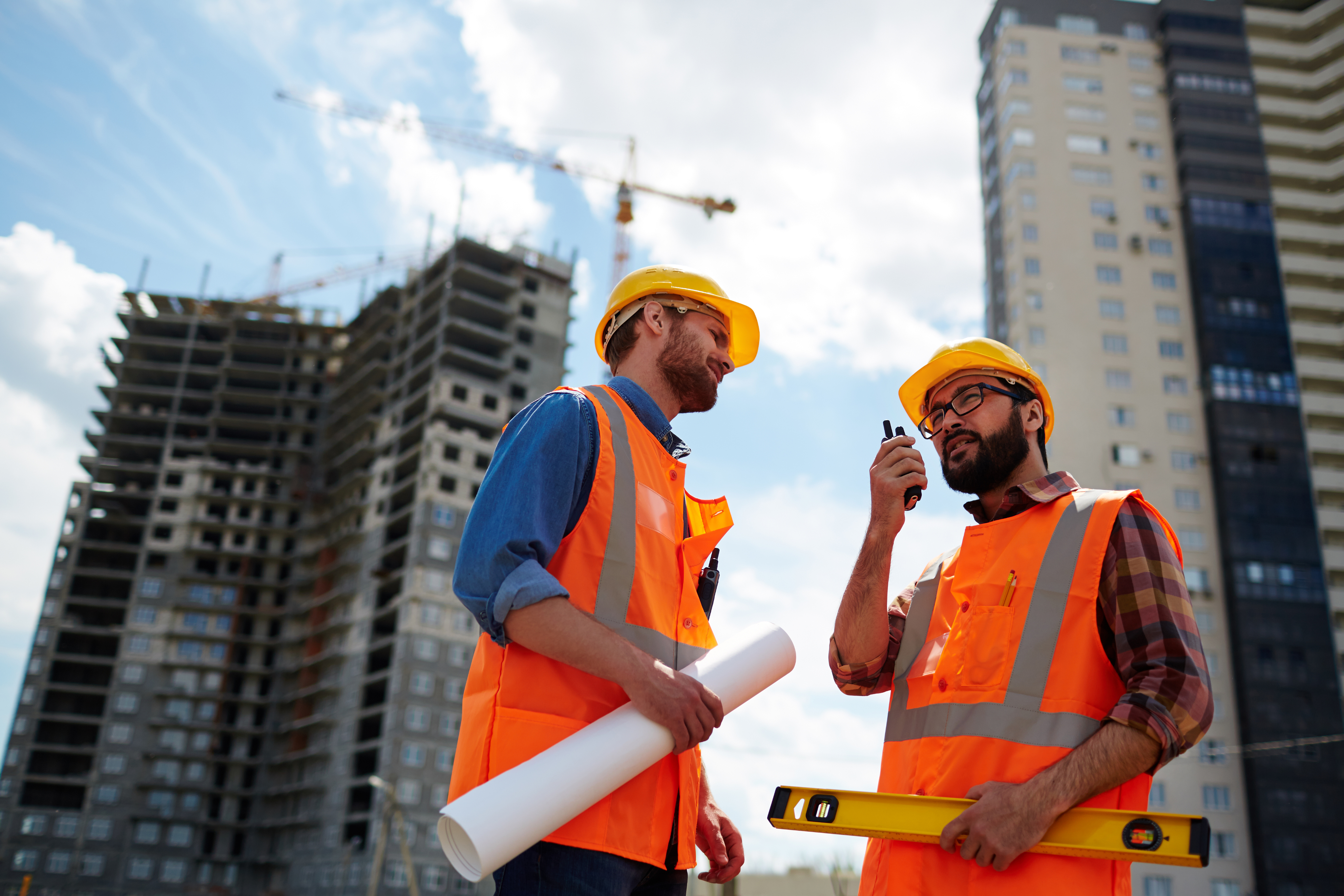 Two construction workers in safety gear discuss at a building site. One holds plans, the other uses a walkie-talkie. Tall buildings tower in the background.