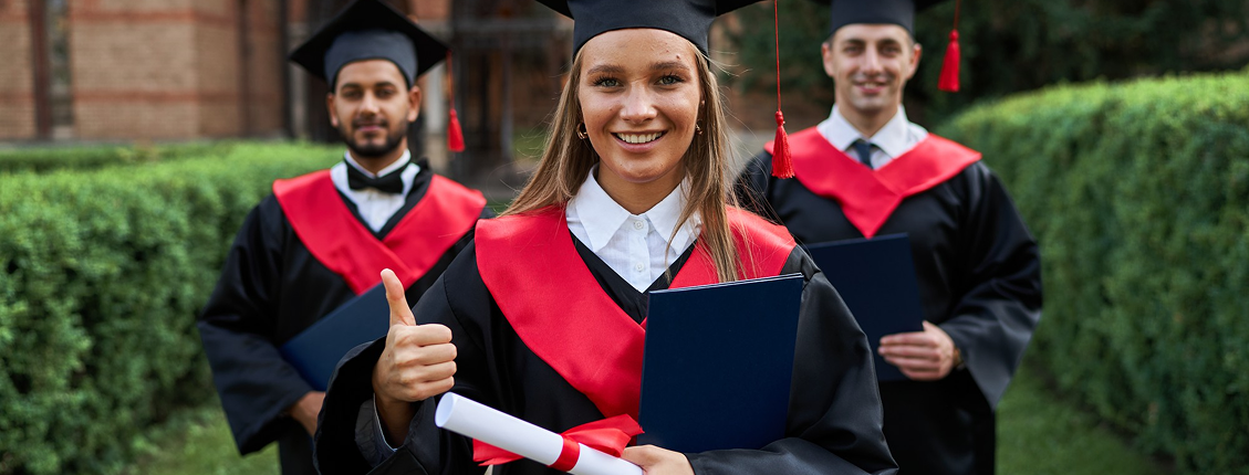 Three graduates in black gowns and red sashes stand outdoors, smiling. The woman in front holds a diploma and gives a thumbs-up, radiating excitement.