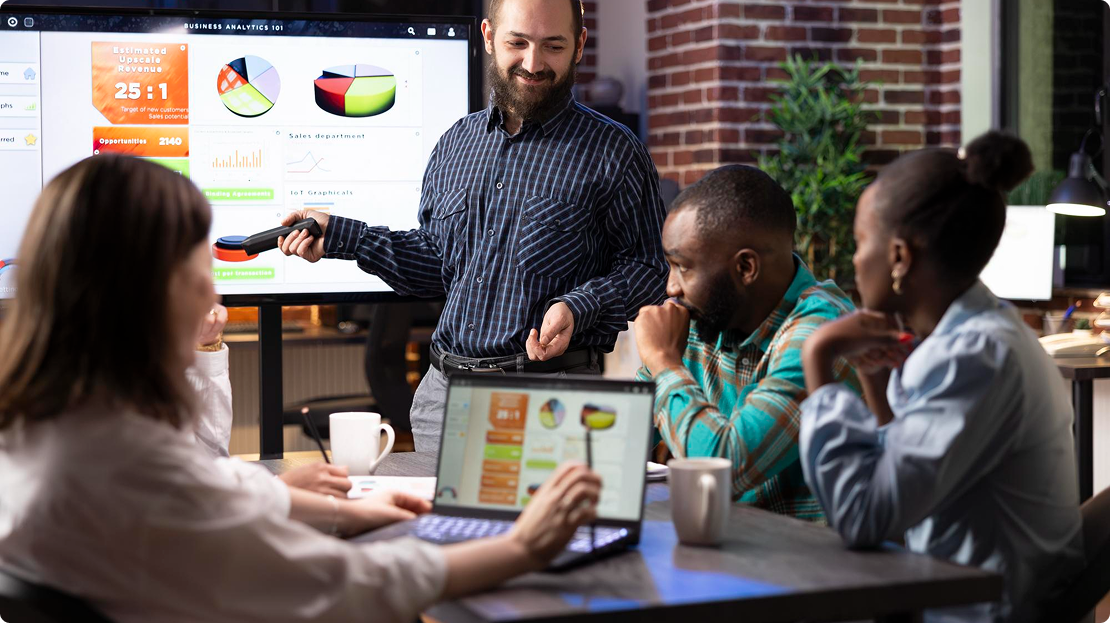 A man presents charts on a screen to three colleagues at a table with laptops. The atmosphere is engaged and collaborative in an office setting.