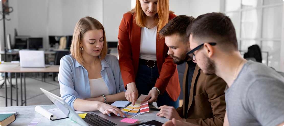 A group of four people collaborates at a desk, analyzing color swatches and sticky notes. The setting is a modern office, suggesting teamwork and creativity.