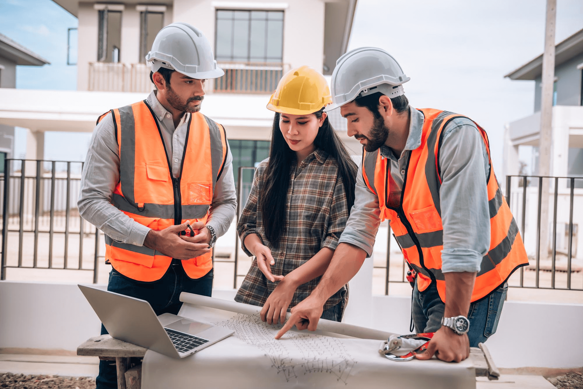 Three construction workers in safety vests and helmets discuss architectural plans at a site, displaying focus and teamwork. A laptop is on the table.