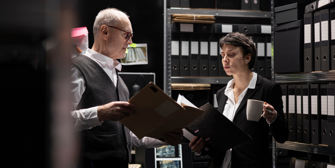 An older man and a younger woman, both in business attire, review documents in a dimly lit archive room filled with shelves of binders, conveying focus.