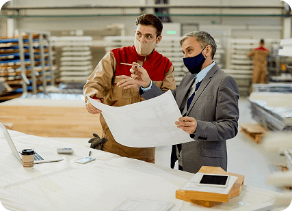 Two men wearing masks work together in a factory setting, focused on their tasks amidst machinery and equipment.