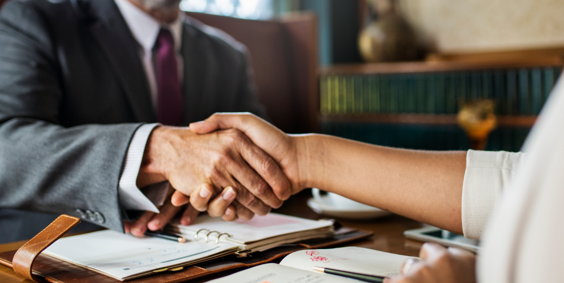 Two people shake hands over a table with open planners, conveying a professional agreement. The setting is business-like and cordial.