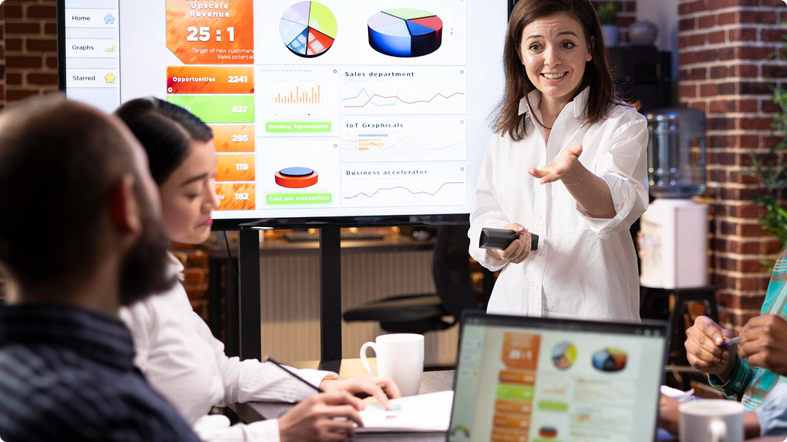 A woman presents data with charts on a screen in a meeting room. Three colleagues listen attentively. The scene conveys collaboration and engagement.