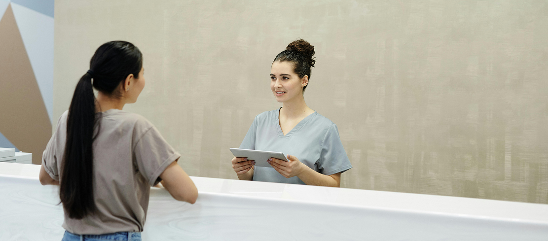 A receptionist in gray scrubs holds a tablet and smiles at a woman with a ponytail across a white counter, conveying a friendly, professional atmosphere.