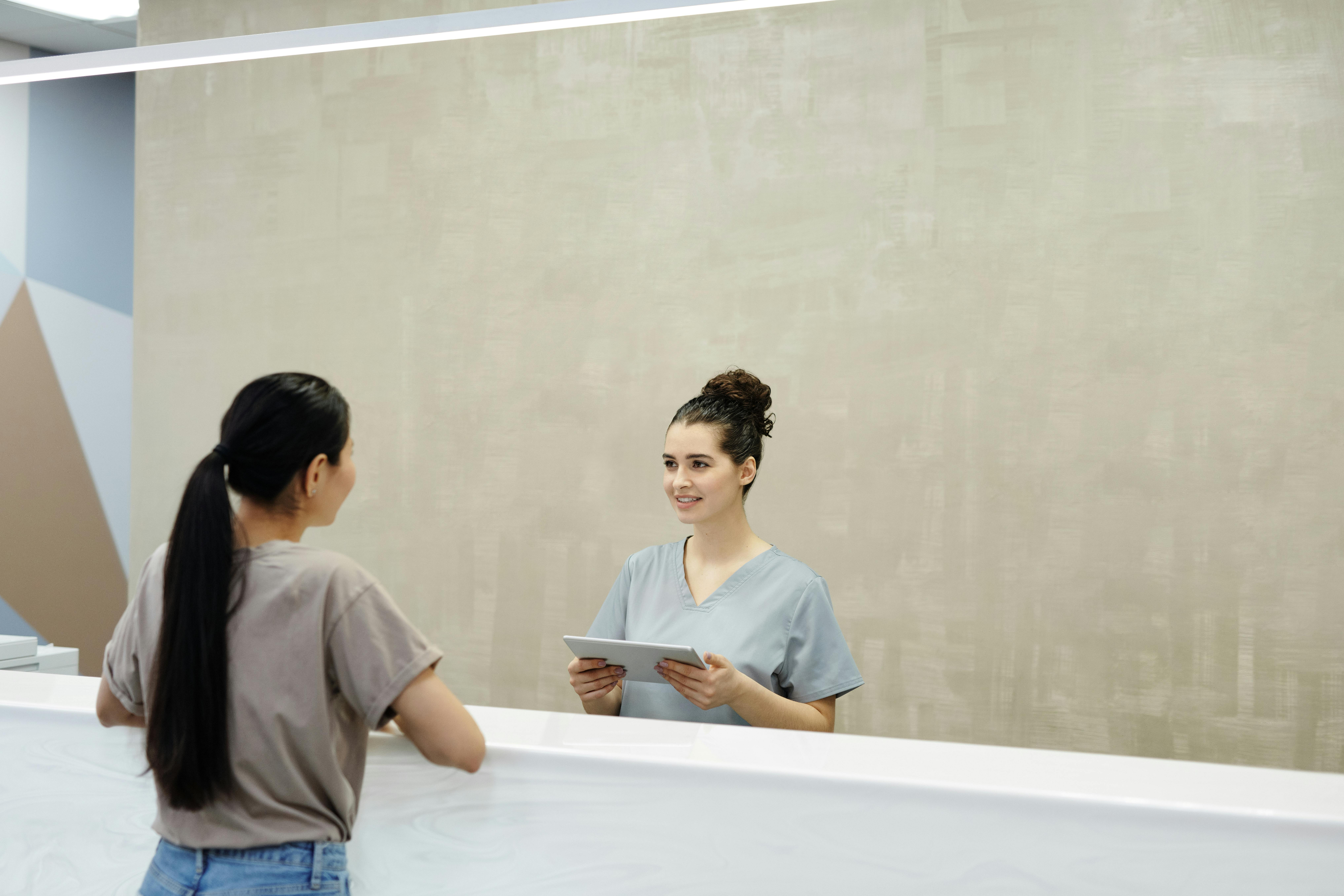 A receptionist in gray scrubs holds a tablet and smiles at a woman with a ponytail across a white counter, conveying a friendly, professional atmosphere.