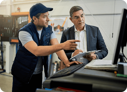 Two men in a factory examining data on a computer screen, surrounded by industrial equipment.
